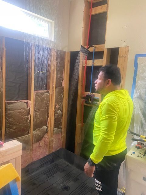 Man in yellow shirt inspects a shower under construction. Exposed studs, showerhead sprays water.