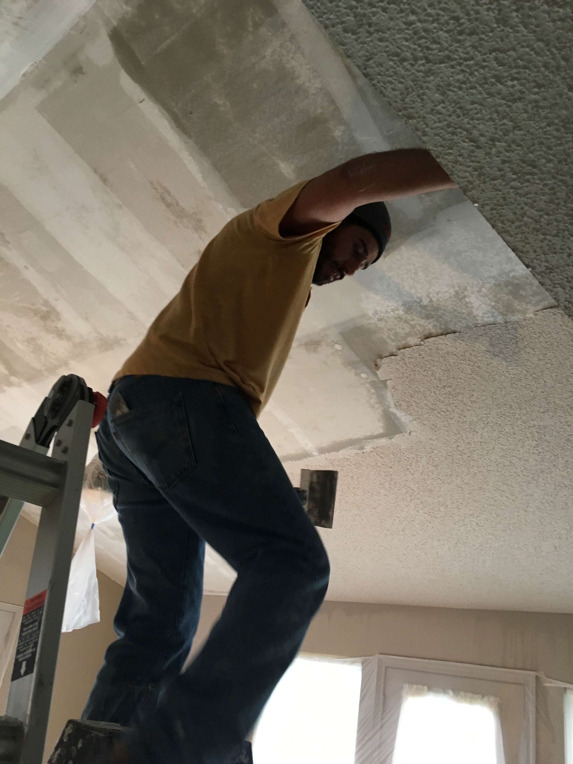 Person on a ladder removing ceiling texture, wearing jeans and a yellow shirt in a room with a window.