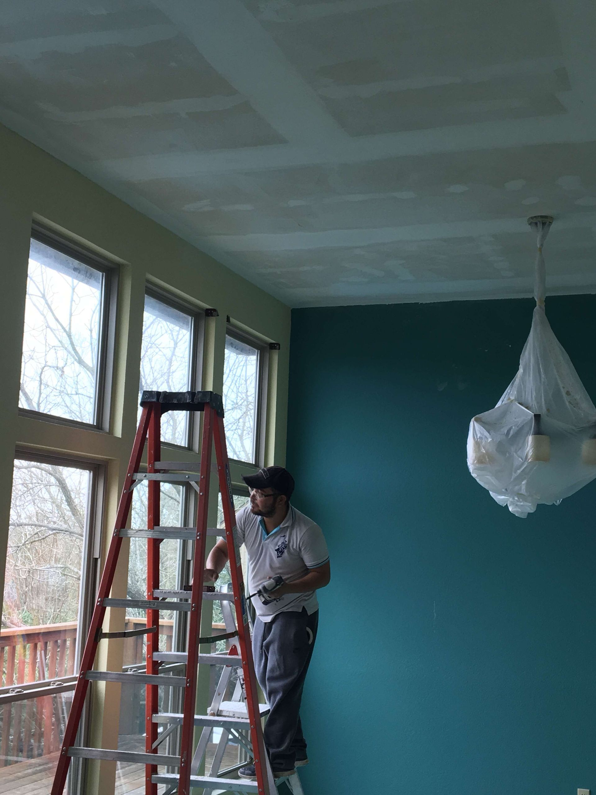 Man on ladder painting ceiling, next to large windows and teal wall.