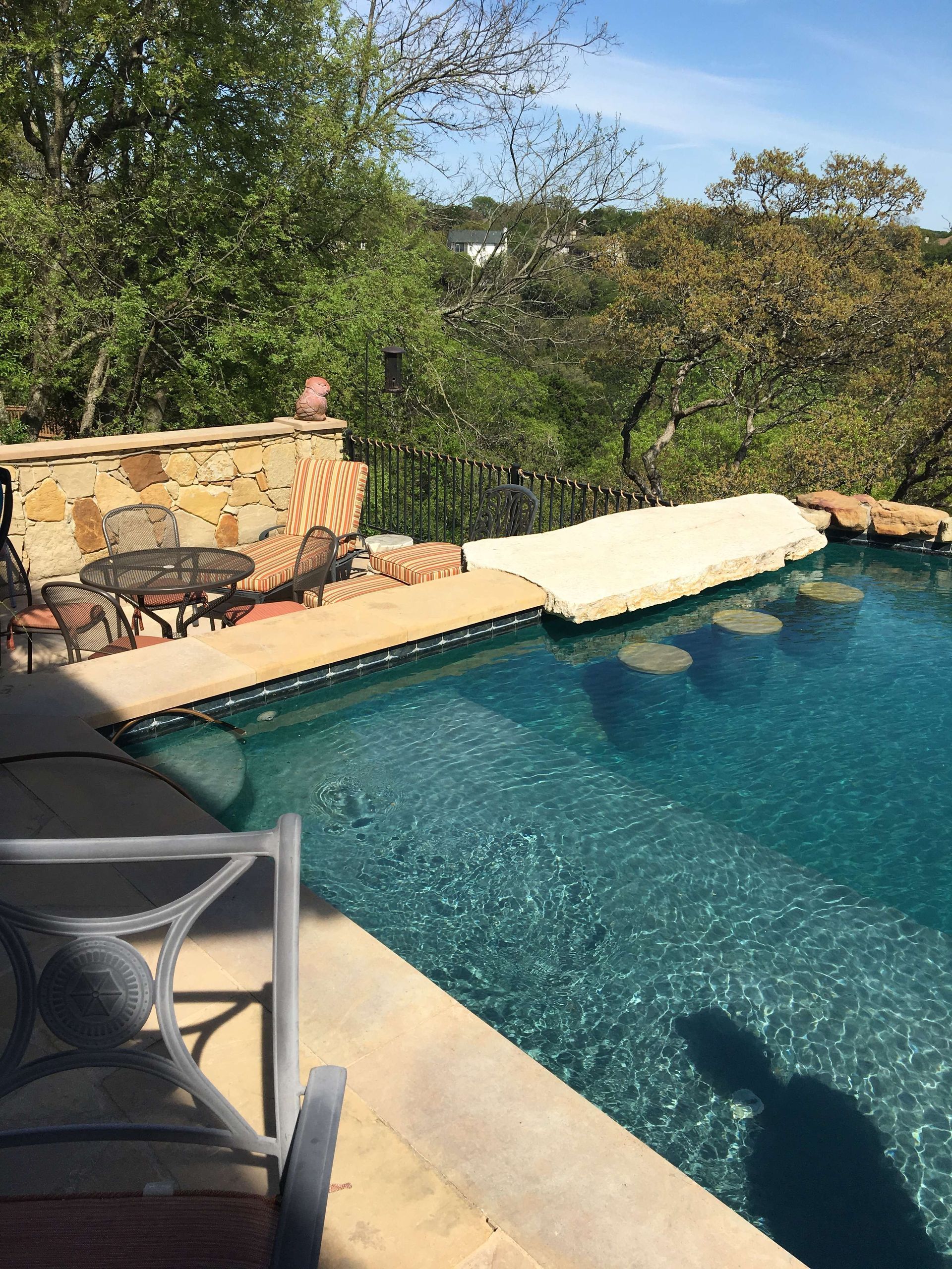 Swimming pool with seating area, stone wall, and trees on a sunny day.