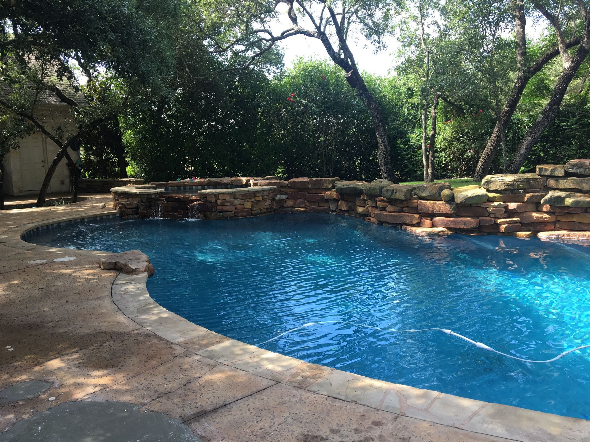 A blue swimming pool with a stone waterfall feature in a lush, green backyard setting.