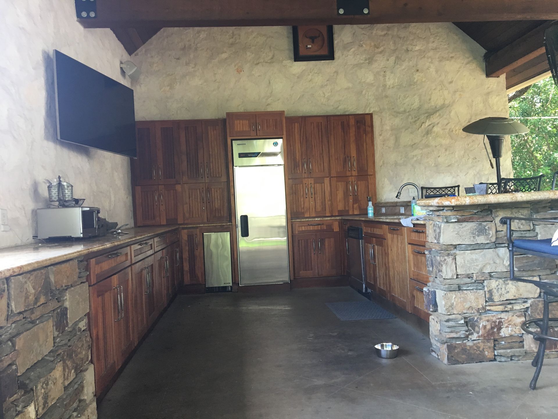 Outdoor kitchen with stone accents, wood cabinets, appliances, and a mounted TV.