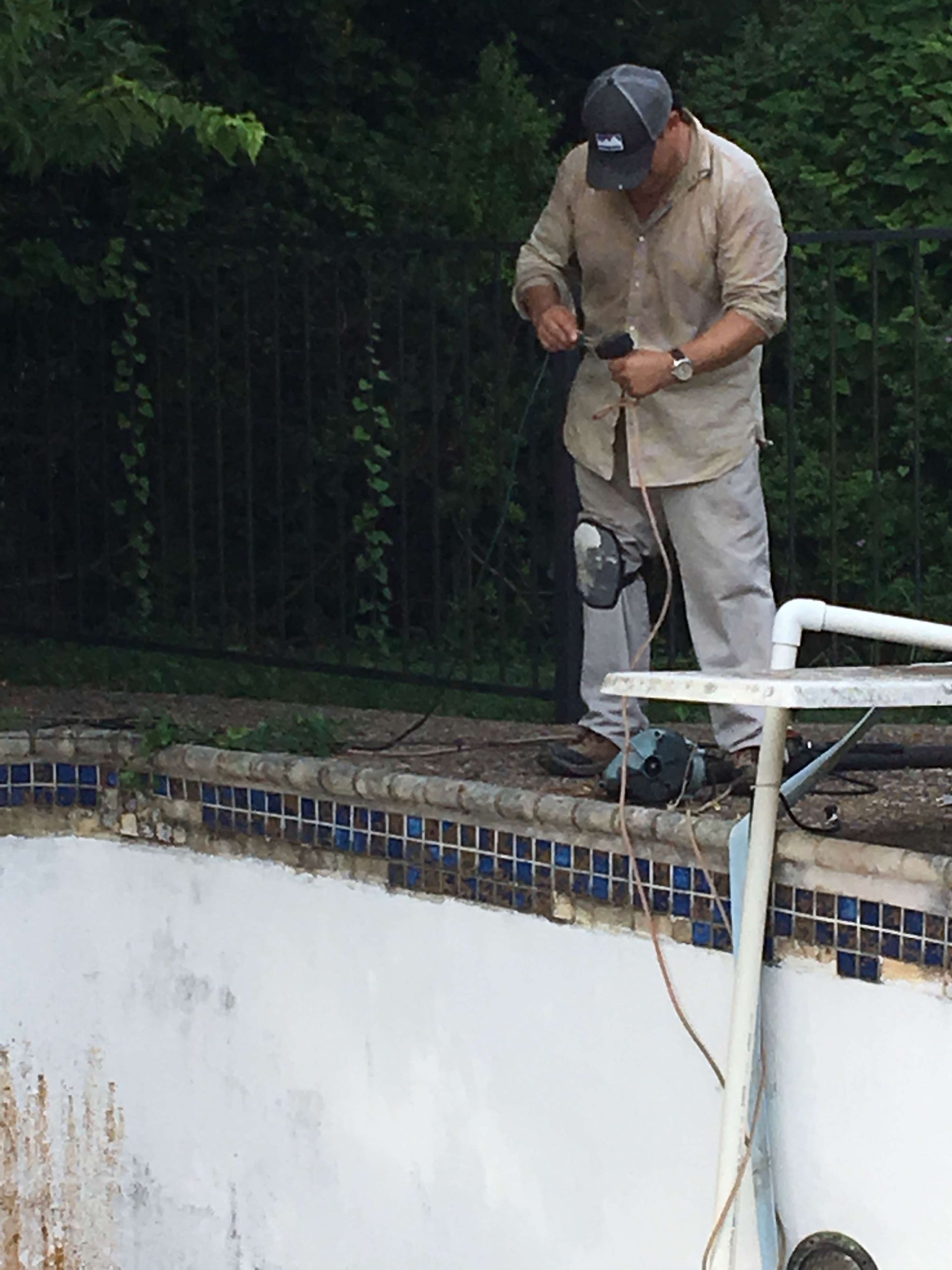 Man working on a pool's edge, using tools. Outdoors, daytime. The pool is empty and dirty.