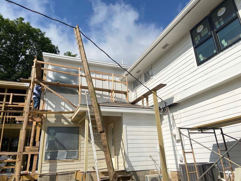 House under construction with wood frame, scaffolding, and siding; worker on the frame against blue sky.
