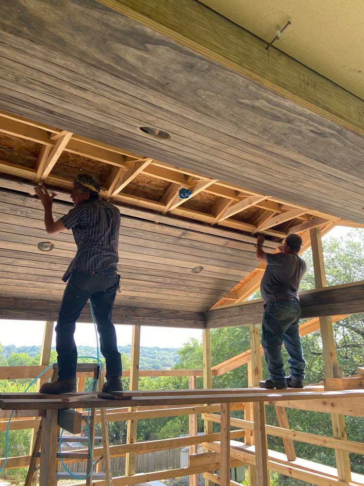 Two construction workers installing ceiling panels on a wooden structure outdoors.