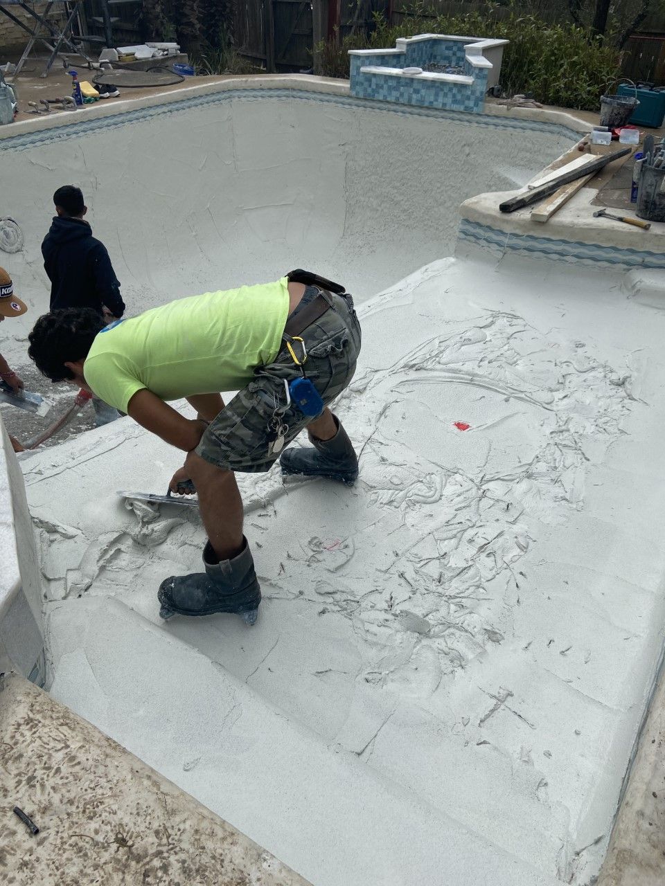 A worker applies coating to a pool, wearing a green shirt and camo shorts.