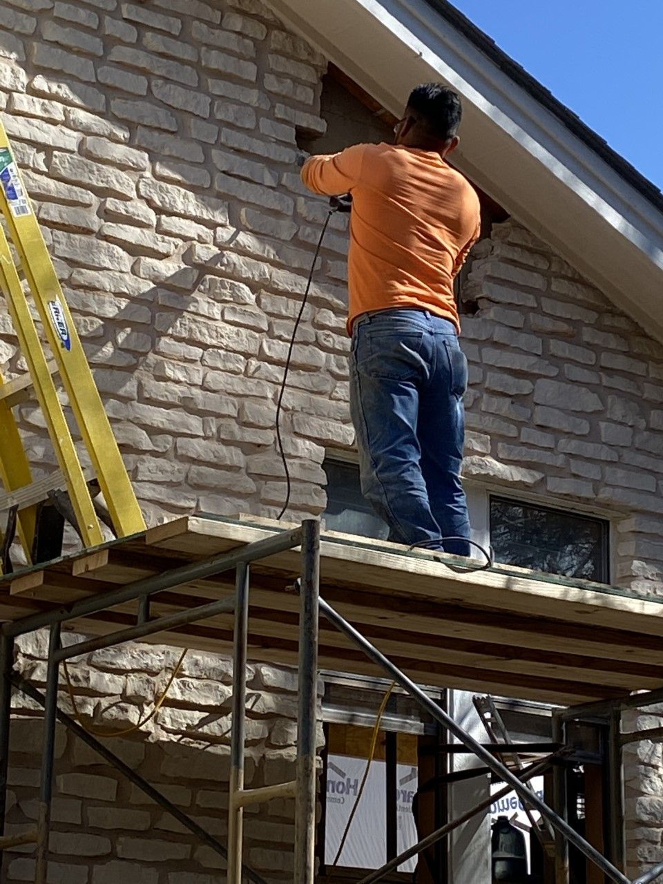 Man in orange shirt on scaffolding works on a stone-clad house exterior under the eaves.