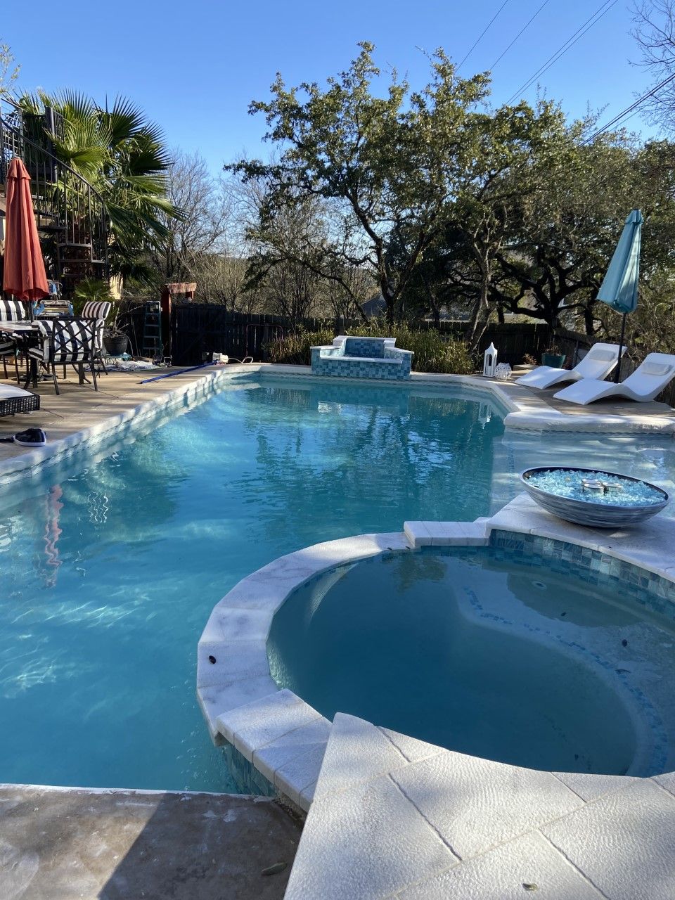 Swimming pool with a connected hot tub, white lounge chairs, and a blue umbrella under a blue sky.