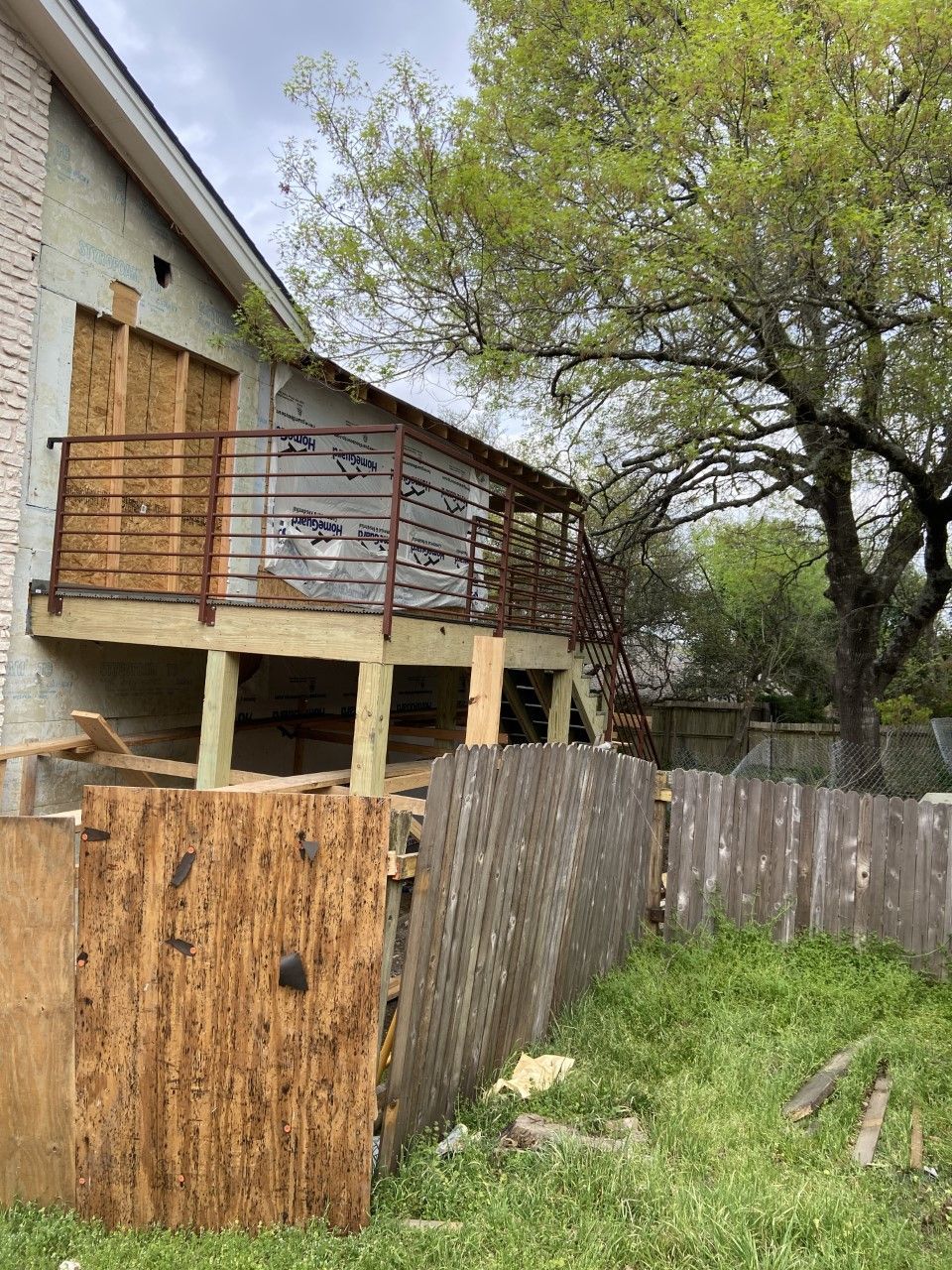 Exterior view of a house undergoing construction, with boarded windows, deck, and a wooden fence.