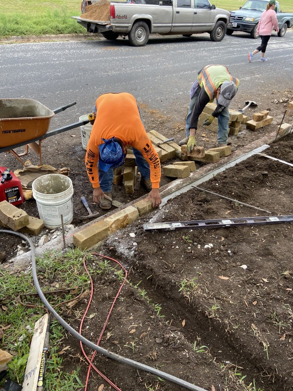 Two construction workers laying bricks outdoors, one bent over, other using a trowel. Orange shirt, safety vest, dirt ground.