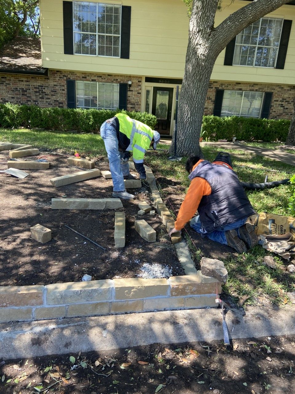 Construction workers building a brick border in a yard near a beige house and tree.