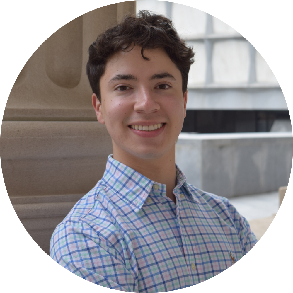 A young man in a blue sweater is smiling in front of a building with columns.