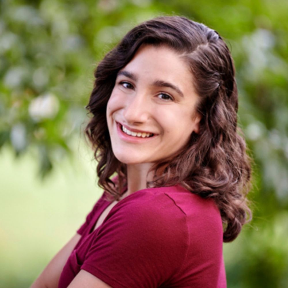 A young woman with curly hair is smiling for the camera while wearing a white t-shirt.