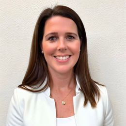 A woman wearing a white jacket and a necklace is smiling for the camera.