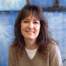 A woman in a brown sweater smiles in front of a blue wall