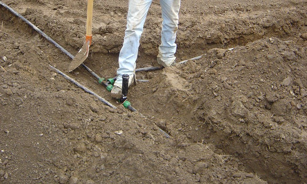le gambe di una persona, una pala in un terreno e dei tubi di un impianto di irrigazione