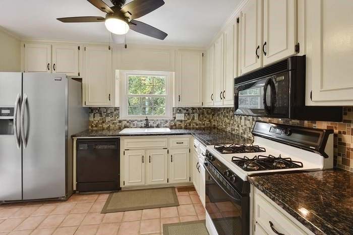 Kitchen with off-white cabinets, dark countertops, stainless steel fridge, black appliances, and brown-tiled backsplash.