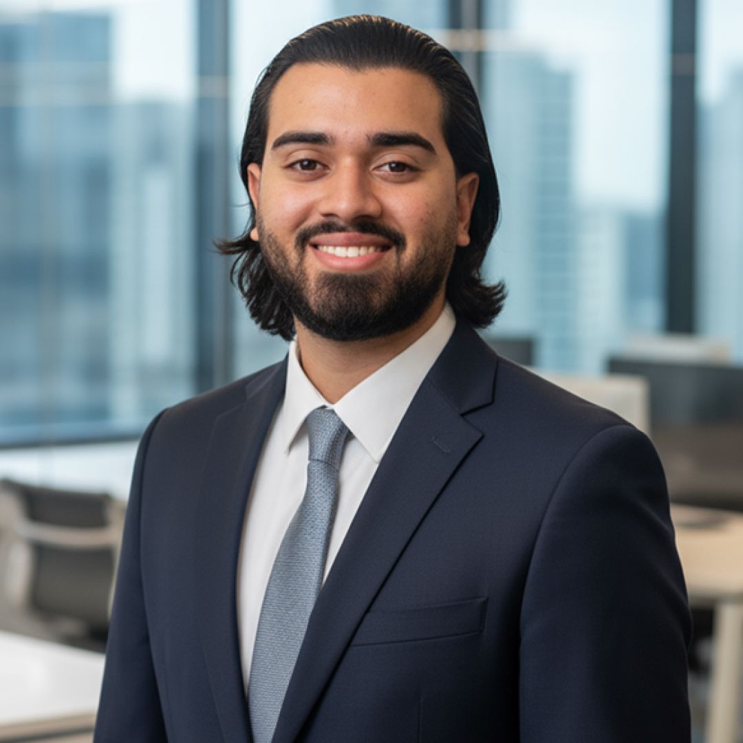 Man in navy suit, tie, smiling in an office setting with a city view.