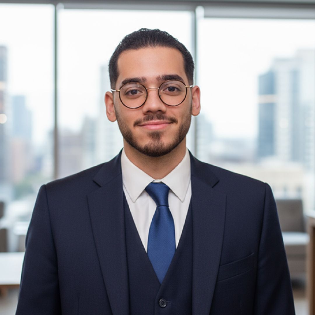 Man in a navy suit and tie, wearing glasses, standing in front of a window with a city view.
