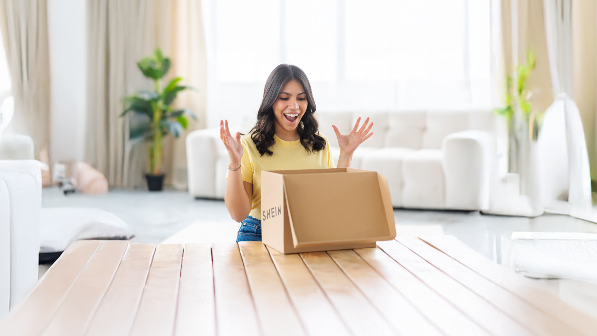 Woman excitedly opens a cardboard box on a table in a bright living room.