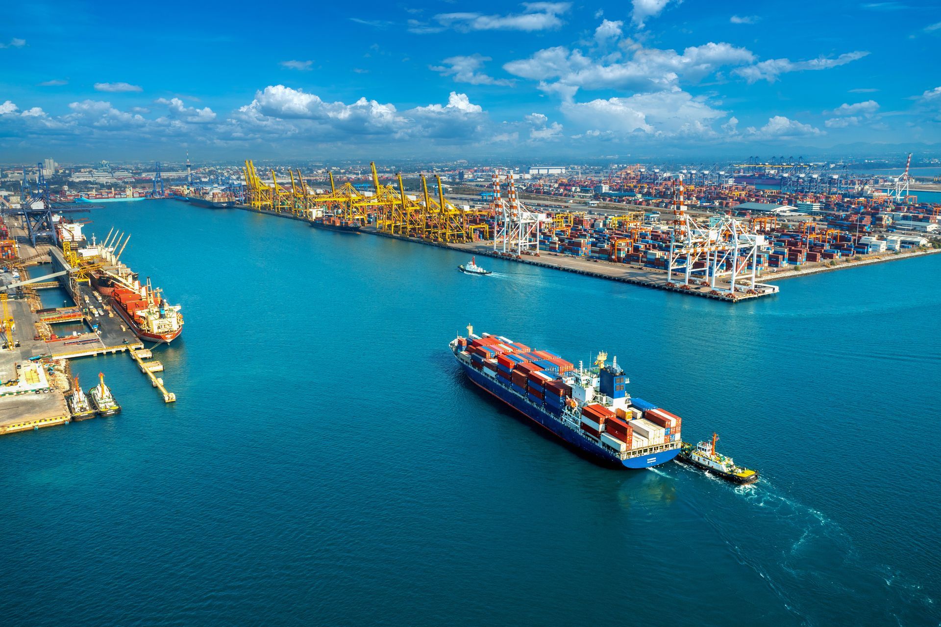 Aerial view of a busy port with container ships, cranes, and cargo. Blue water, sunny day.