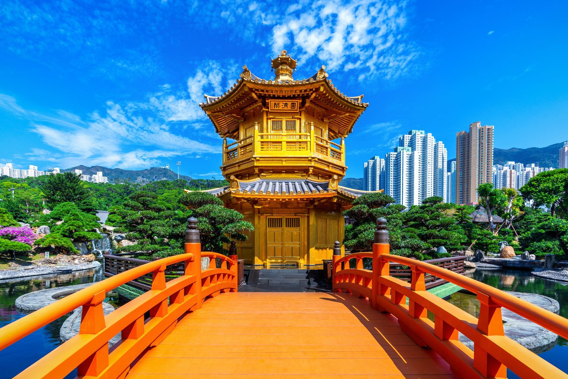 Golden pavilion with orange bridge over water in a tranquil garden, city skyline in the background.