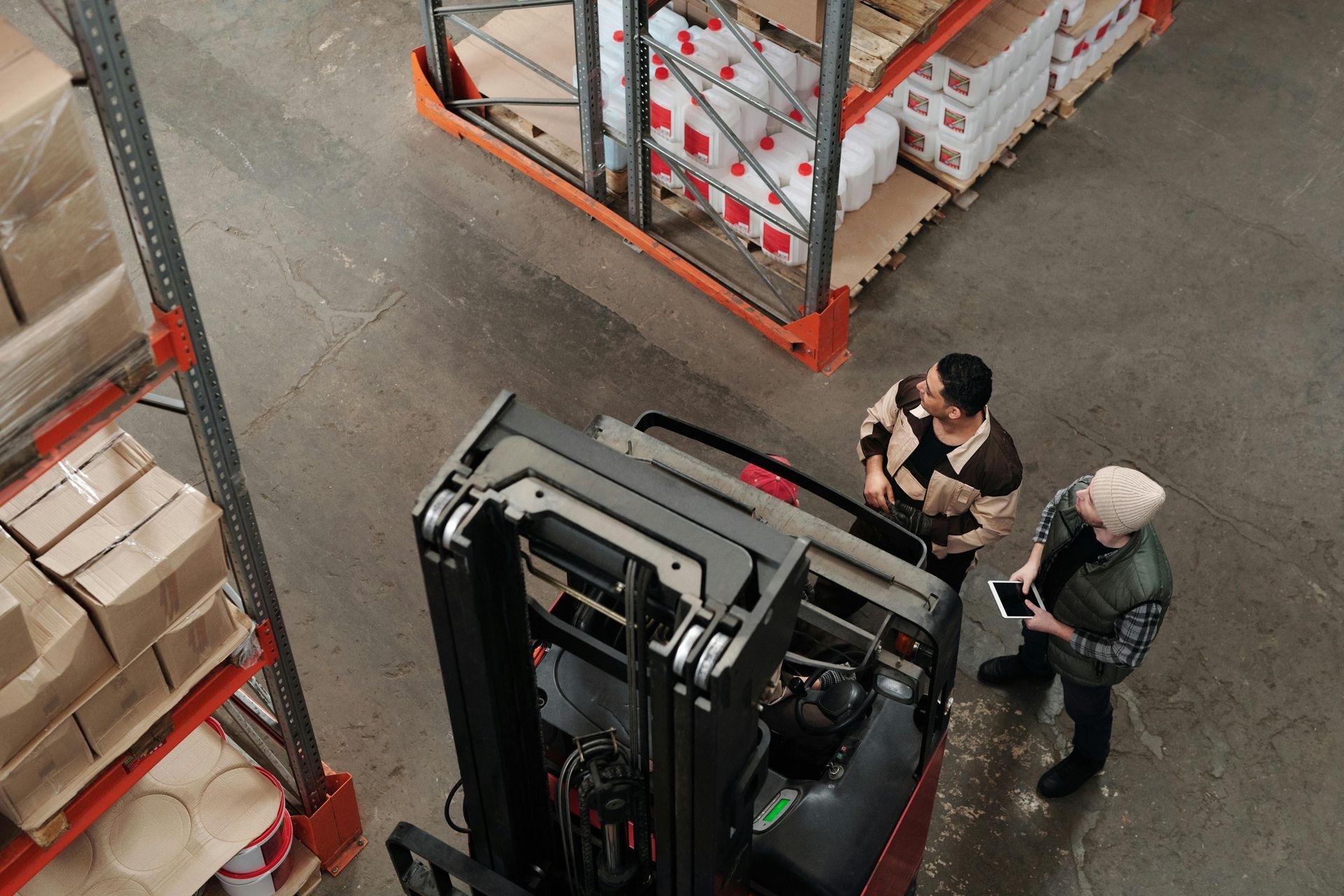 Two people near forklift in warehouse; reviewing tablet, shelves with boxes.