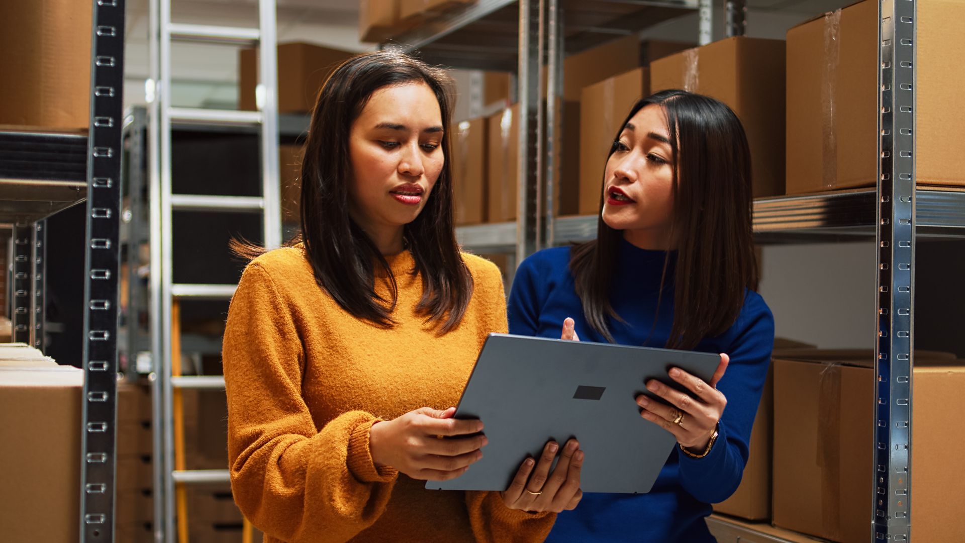 Two women in a warehouse discuss inventory, one pointing at a tablet, surrounded by stacked boxes.