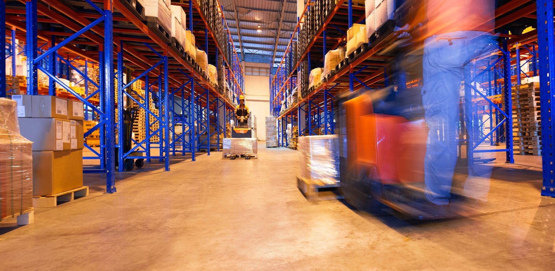 Warehouse interior with forklifts moving goods along aisles lined with shelving.