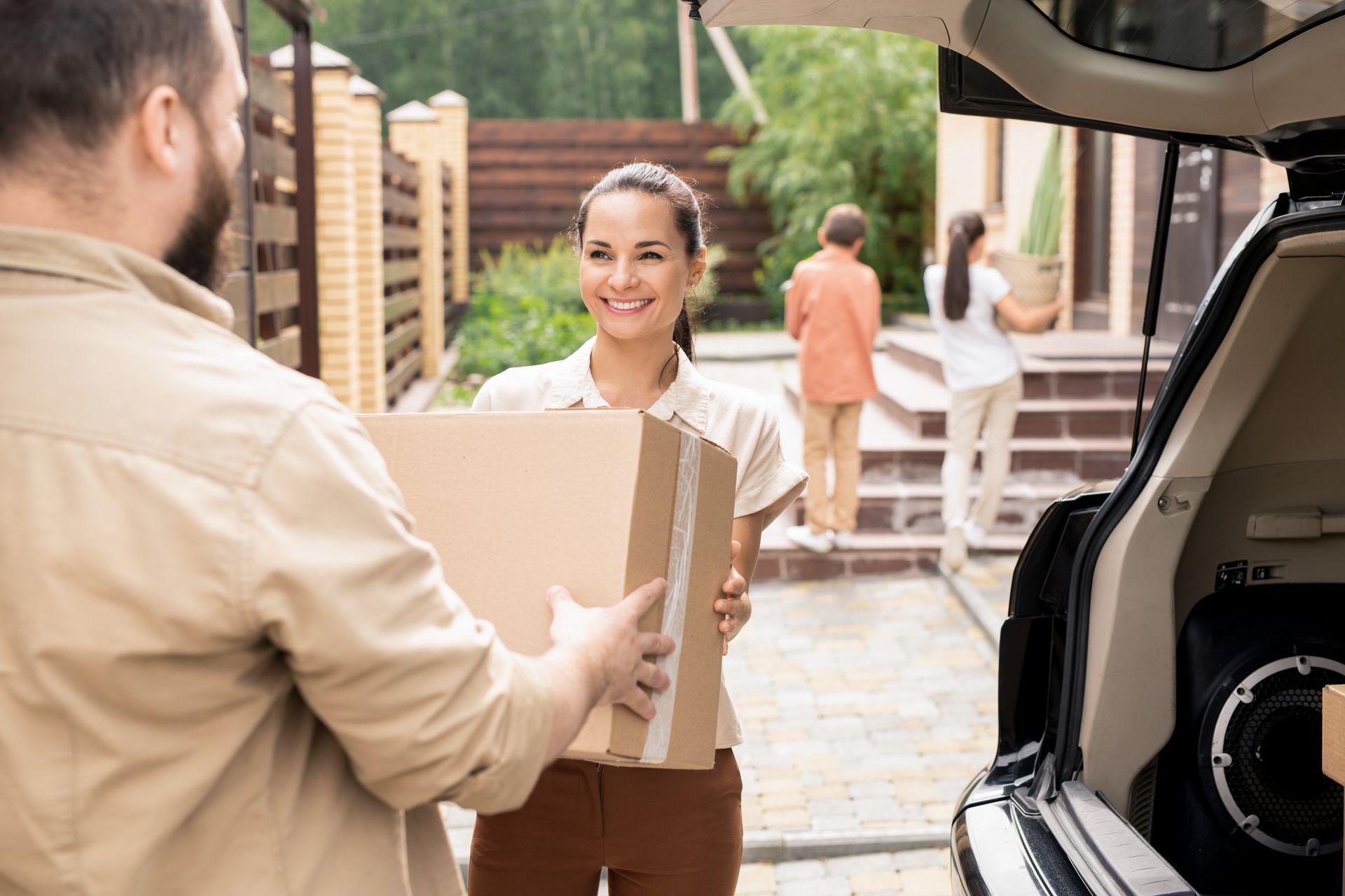 People moving boxes from a car. A woman and man are passing a box; others carry boxes toward a house.
