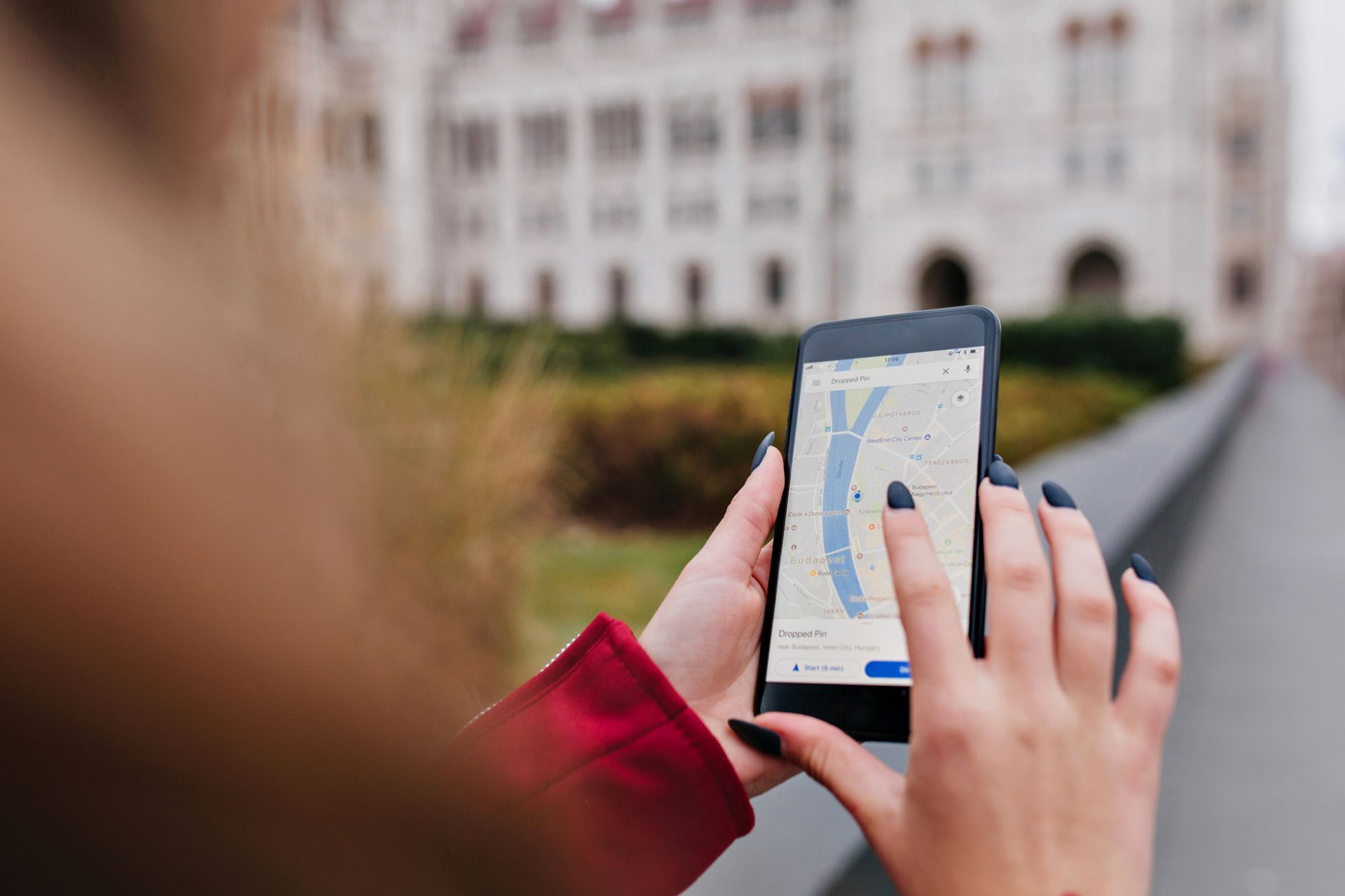 Woman holding smartphone with map app, pointing at screen. Background: building, outdoor.