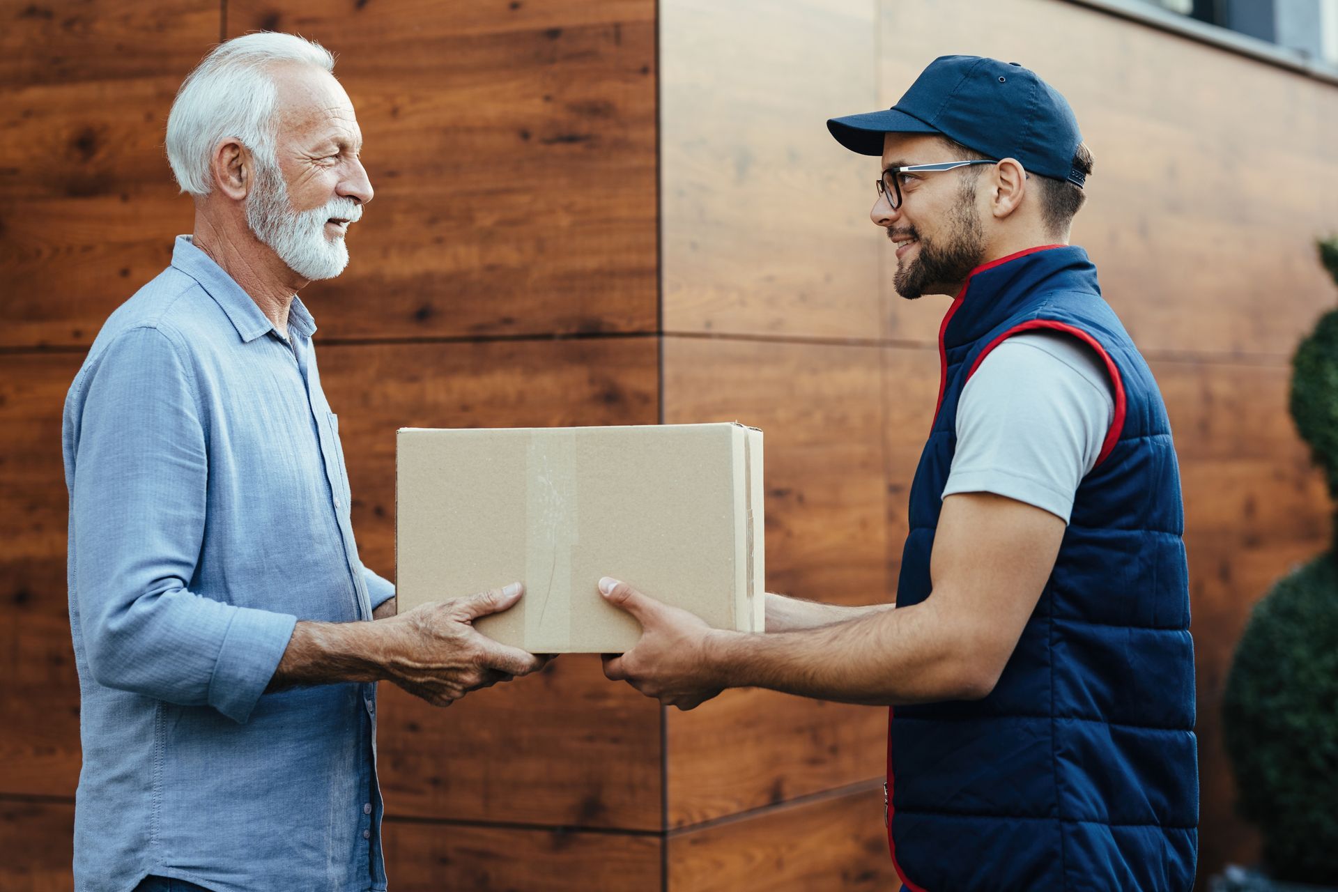 Delivery person handing a cardboard box to an elderly man outside a house. Both smile.