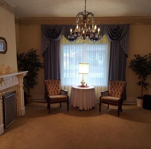 Living room with fireplace, window with draped curtains, chandelier, two armchairs facing a small table with a lamp.