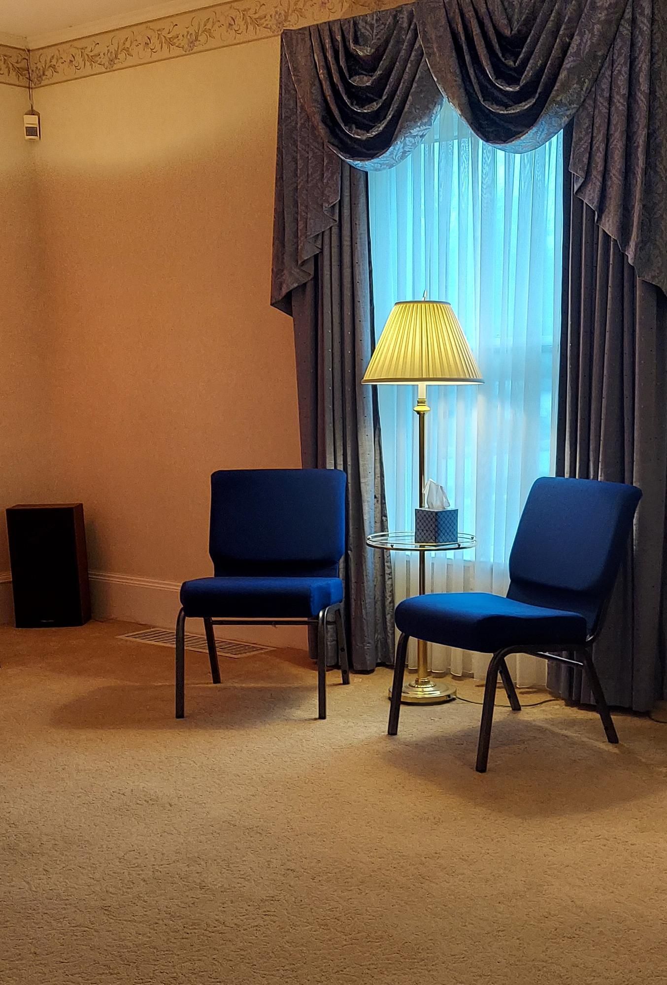 Two blue chairs sit on a carpet, beside a window with blue curtains and a lamp.