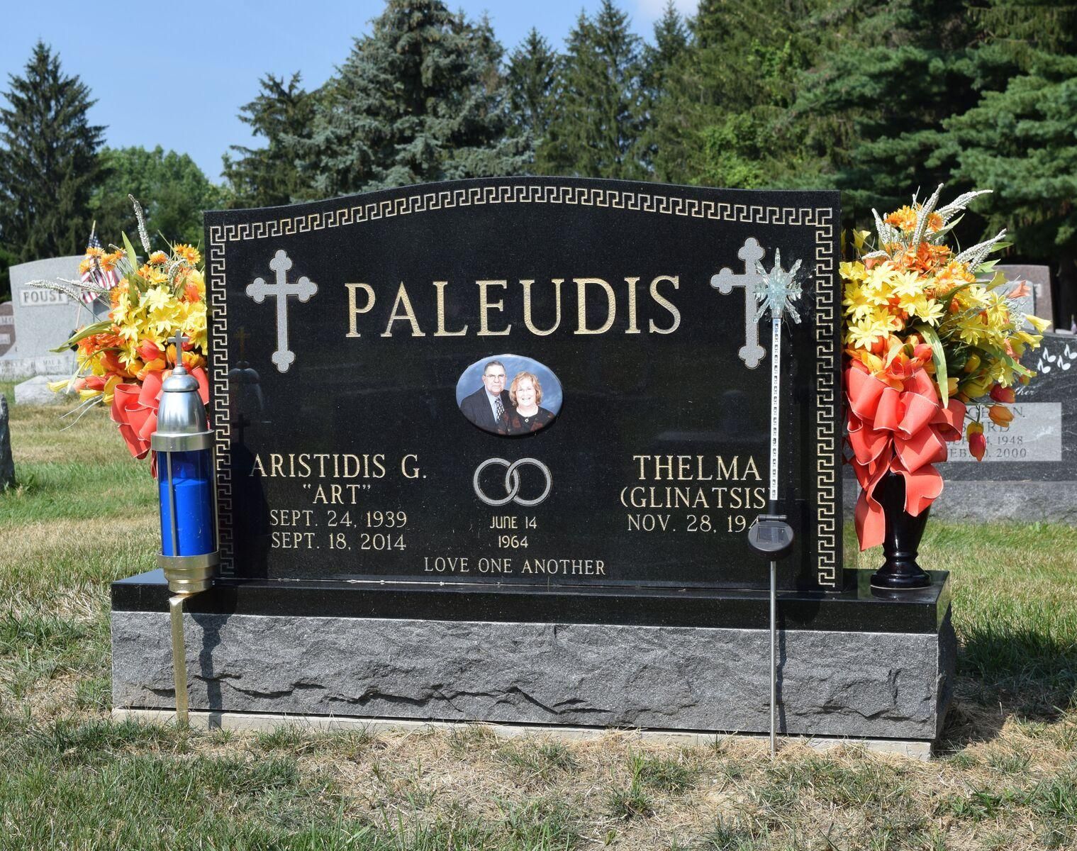Black headstone with gold lettering, photos, cross symbols, and floral arrangements.