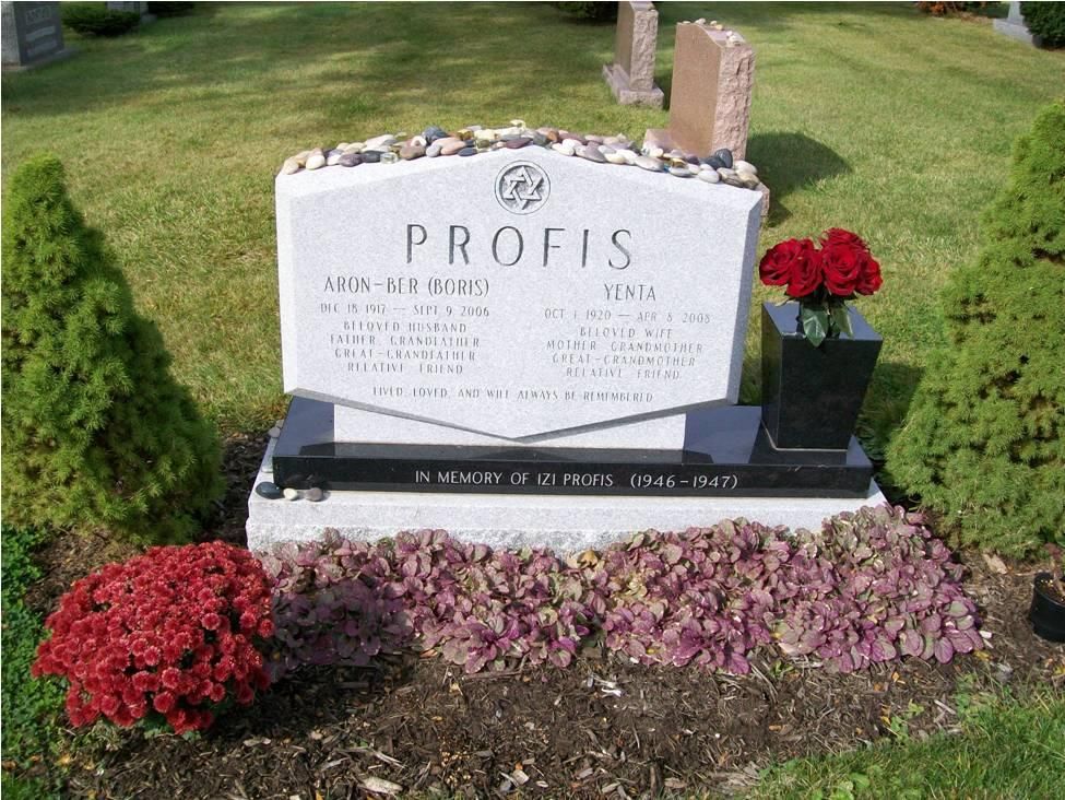 Gravestone for the Profis family in a grassy area, featuring names and dates. Red flowers sit nearby.