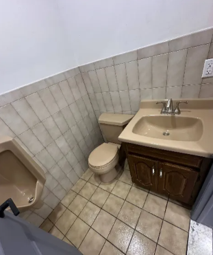 A beige public restroom with a urinal, toilet, and sink with brown cabinet.