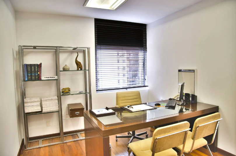 Office with a desk, chairs, shelving unit, and window with blinds. Light wood floors and a tan/brown desk.