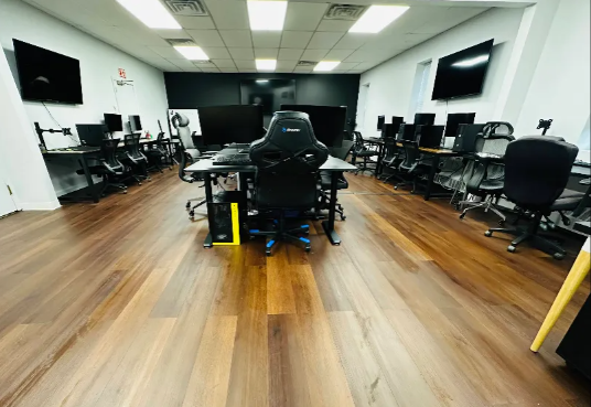 Gaming room with multiple computer stations, black chairs, wooden floors, and overhead lighting.