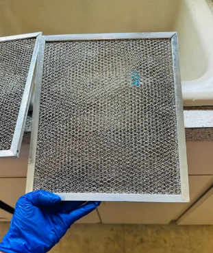 Hand holding a metal range hood filter, dirty with grease, near a sink.