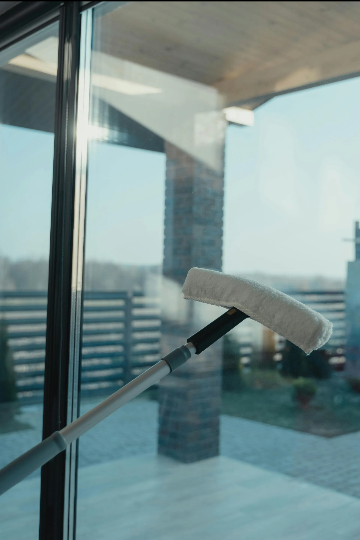 Window being cleaned with a long-handled squeegee; exterior view of house and yard visible through glass.