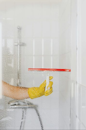 Person in yellow glove uses a red squeegee to clean a glass shower door.