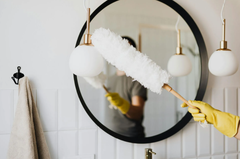 Person in yellow gloves dusting a round mirror in a white bathroom with globe lights.