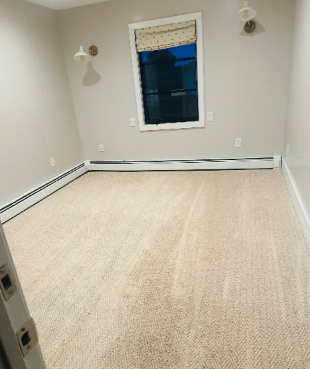 Empty room with beige carpet, off-white walls, small window with shade, and baseboard heaters.