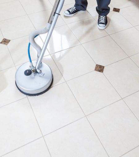 Person cleaning tile floor with a rotary cleaning machine, standing in a room.