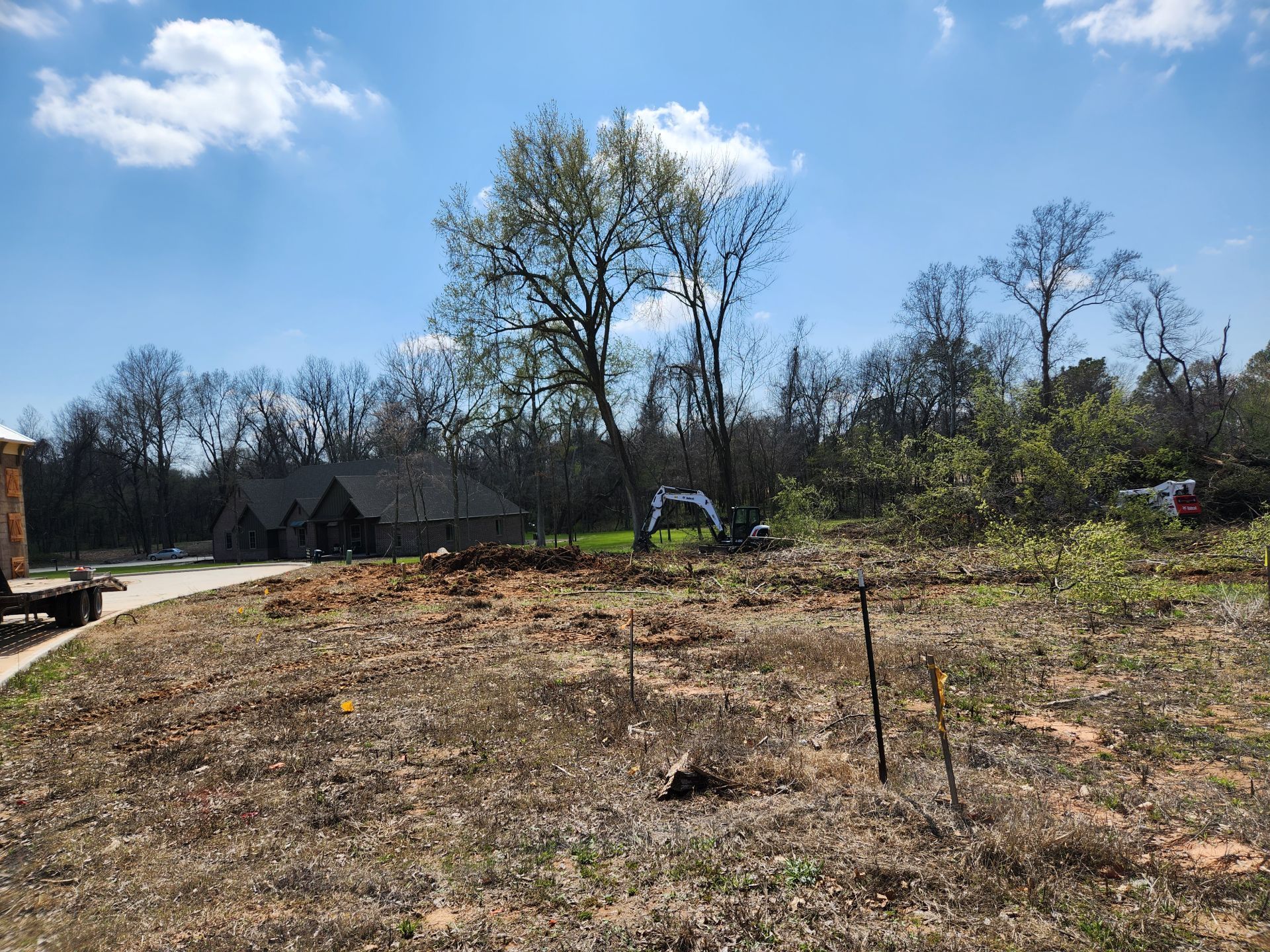 Construction site with dirt and trees, equipment in the distance, and a house in the background.
