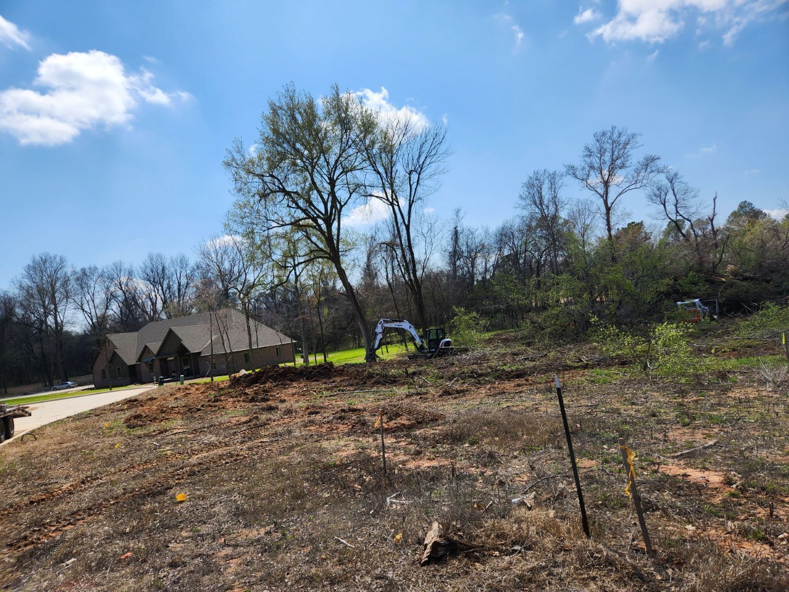 Excavator on a hillside clearing land near a house under a blue sky.