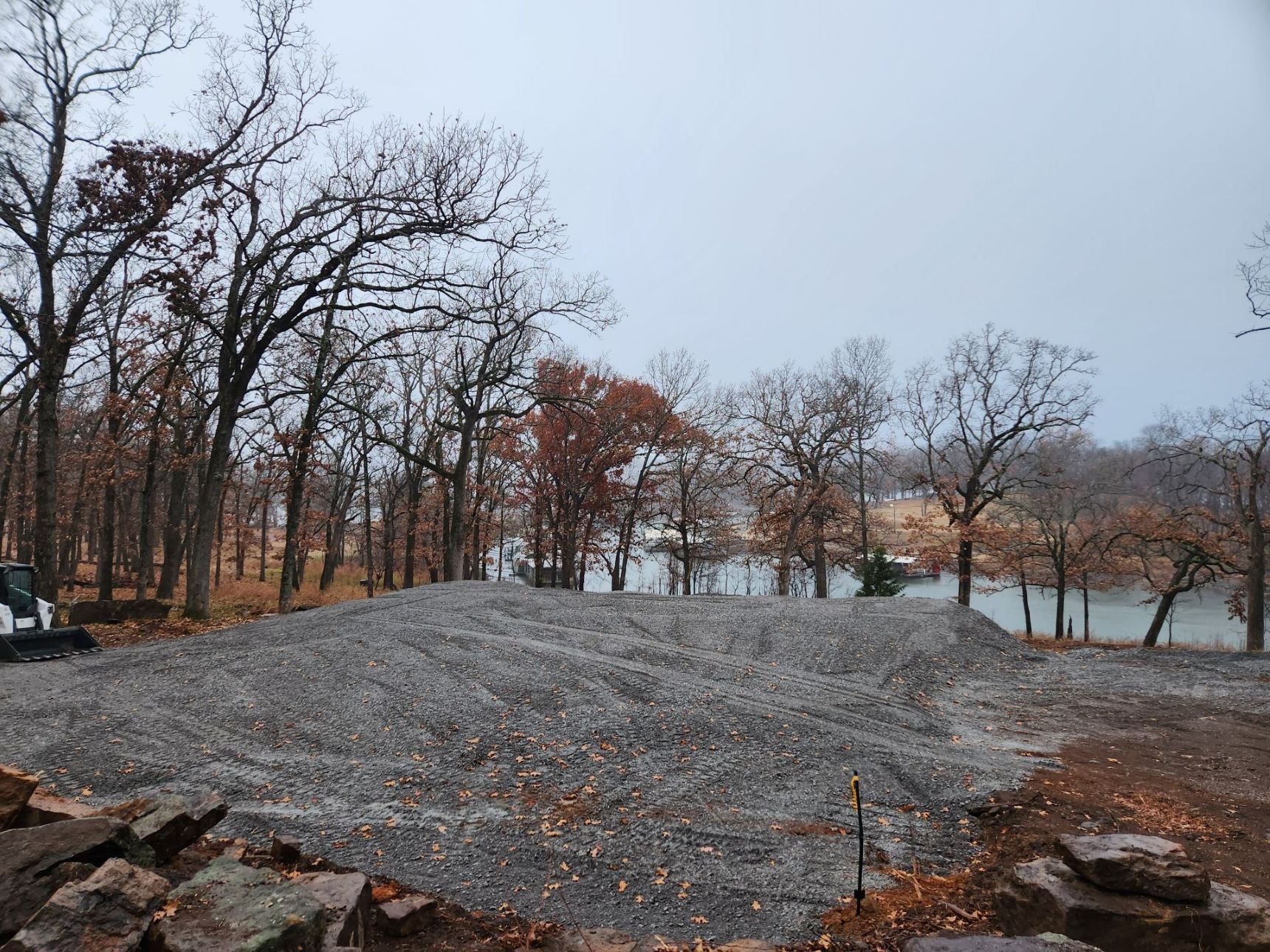 Pile of gray gravel in front of trees and a lake, under an overcast sky.