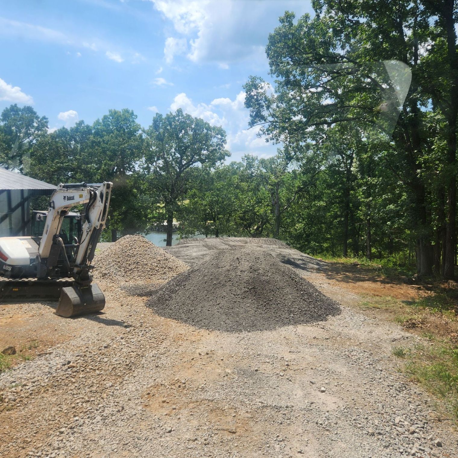 Excavator on a gravel road next to piles of gravel, wooded area in the background, blue sky.
