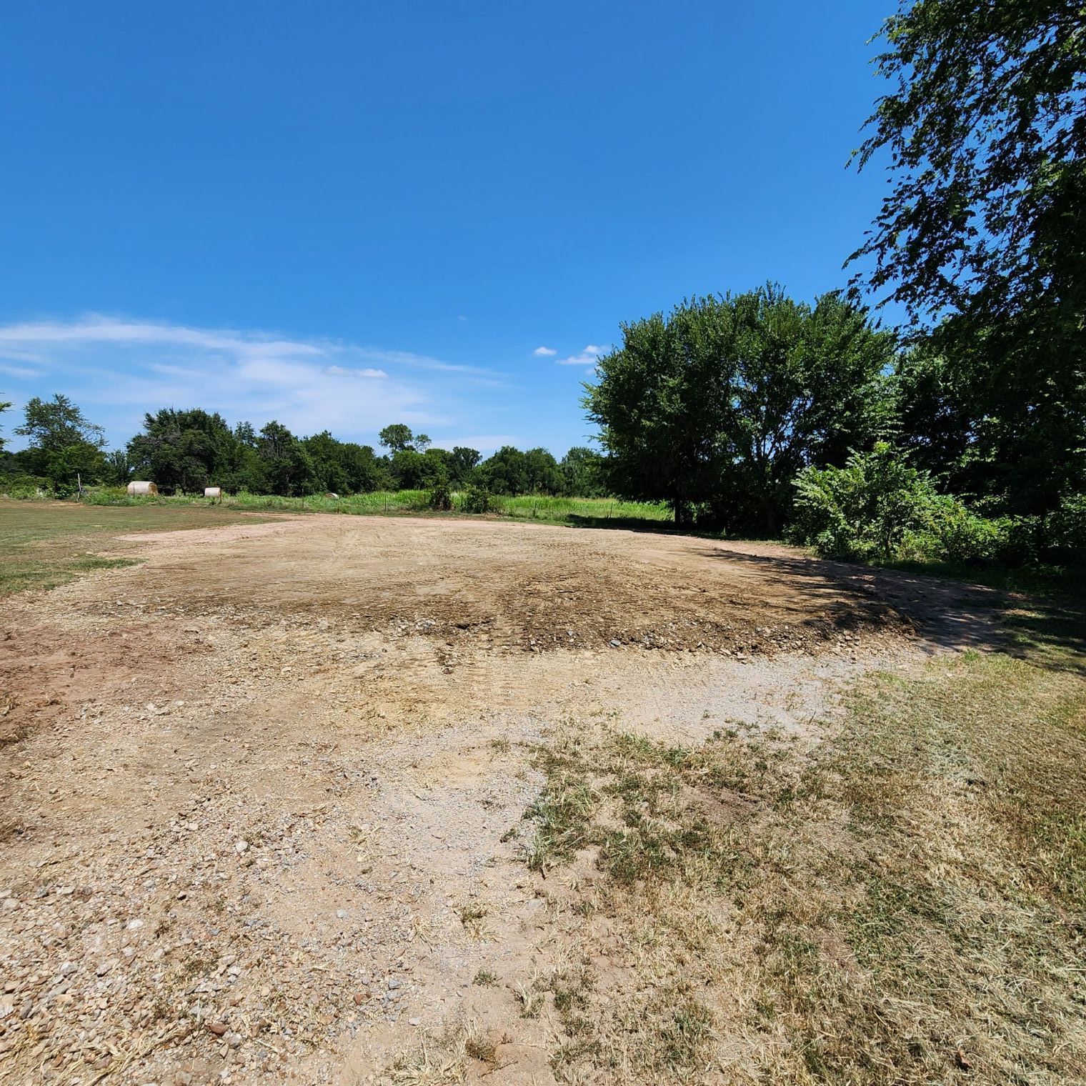 Dirt and gravel lot under a blue sky, surrounded by trees and grass.